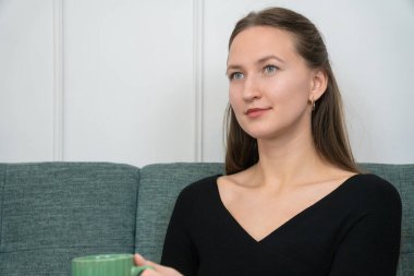 Siyah bluzlu sakin bir kadın, yeşil bir kupa tutan, deniz mavisi bir kanepede rahat bir şekilde oturur ve düşünceli bir şekilde uzaktan bakar, huzurlu bir anın tadını çıkarır.