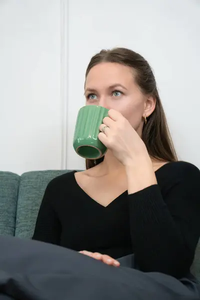 Genç bir kadın rahat bir kanepede rahatlatıcı bir anın tadını çıkarıyor, evinin rahat atmosferinde şık yeşil bir bardaktan ılık bir içeceğin tadını çıkarıyor. Dikey fotoğraf