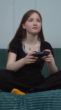 Genç bir kadın oturma odasındaki kanepede bağdaş kurarak oturuyor, kablosuz kontrolörle video oyunu oynarken gündelik kıyafetlerini giyiyor, oyun sırasında duygusal tepkilerini gösteriyor.