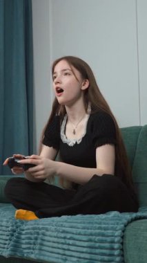 Genç kız rahat koltukta dinleniyor. Derin bir konsantrasyonla kablosuz kontrolörle video oyunları oynuyor. Ev ortamında rahat bir şekilde dikey video seyrediyor.