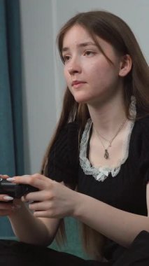 Genç kız rahat koltukta dinleniyor. Derin bir konsantrasyonla kablosuz kontrolörle video oyunları oynuyor. Ev ortamında rahat bir şekilde dikey video seyrediyor.