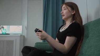 Genç kız rahat koltukta dinleniyor, yoğun konsantrasyonu var. Kablosuz kontrolörle video oyunları oynuyor, evinde rahat bir yaşam sürüyor.