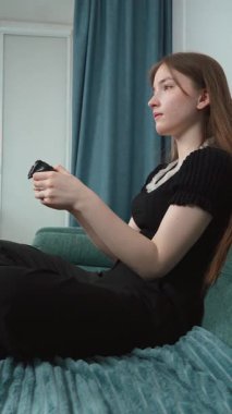 Genç kız rahat koltukta dinleniyor. Derin bir konsantrasyonla kablosuz kontrolörle video oyunları oynuyor. Ev ortamında rahat bir şekilde dikey video seyrediyor.