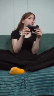 Kız Oyuncu Rahat Ev Ortamı dikey videosundaki video oyununun tadını çıkarırken Kanepede bacak bacak bacak üstüne atarak Kablosuz Oyun Kontrolörü