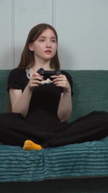 Kız Oyuncu Rahat Ev Ortamı dikey videosundaki video oyununun tadını çıkarırken Kanepede bacak bacak bacak üstüne atarak Kablosuz Oyun Kontrolörü