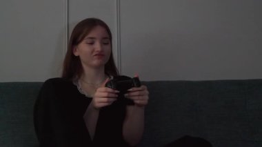 Genç kız kanepede bağdaş kurup video oyunları oynuyor. Yoğun duygusal tepkiler gösteriyor. Yoğunlaştırılmış konsantrasyonla dijital oyun oynuyor.