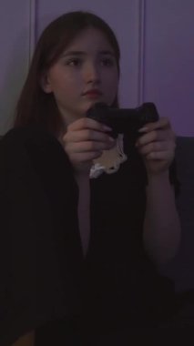 Siyah elbiseli ve sarı çoraplı ergen kız karanlıkta oturma odasındaki turkuaz kanepede otururken kumandayla video oyunu oynuyor. dikey video