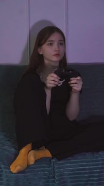 Siyah elbiseli ve sarı çoraplı ergen kız karanlıkta oturma odasındaki turkuaz kanepede otururken kumandayla video oyunu oynuyor. dikey video