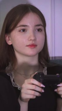 Siyah elbiseli ve sarı çoraplı ergen kız karanlıkta oturma odasındaki turkuaz kanepede otururken kumandayla video oyunu oynuyor. dikey video