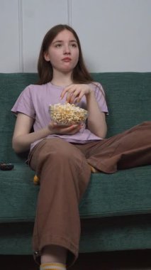 Genç kız rahat bir şekilde yeşil koltukta oturmuş, evde dinlenirken patlamış mısırın tadını çıkarıyor ve dikey televizyon seyrediyor.