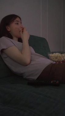 Genç bir kız, evdeki dikey videoda, yumuşak kanepeye uzanıp televizyon seyrediyor ve cam kaseden patlamış mısırın keyfini çıkarıyor.