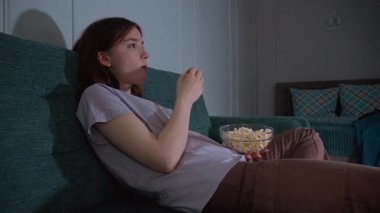 Genç kız yumuşak koltukta dinleniyor, televizyon seyrediyor ve evde boş bir akşamda cam kaseden patlamış mısırın tadını çıkarıyor.
