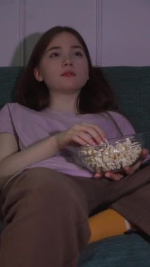 Genç kız kanepede oturup patlamış mısır yiyor ve loş aydınlatılmış oturma odasında film izliyor rahat ve rahatlatıcı bir atmosfer videosu yaratıyor.