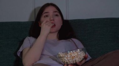 Genç kız kanepede oturmuş patlamış mısır yiyor ve loş ışıklı oturma odasında film seyrediyor rahat ve rahatlatıcı bir atmosfer yaratıyor.
