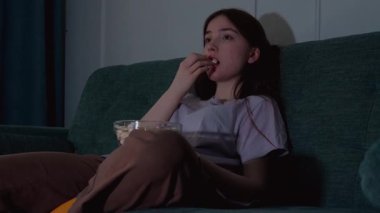 Genç kız kanepede oturmuş patlamış mısır yiyor ve loş ışıklı oturma odasında film seyrediyor rahat ve rahatlatıcı bir atmosfer yaratıyor.