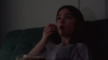 Genç kız kanepeye oturup loş ışıklı odada cam kaseden patlamış mısır yiyerek eğleniyor.