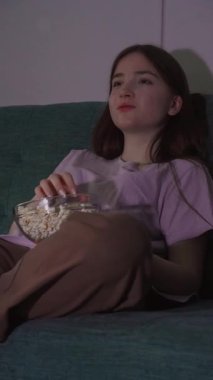 Genç kız kanepeye oturup film izleyerek loş ışıklı oda dikey videosunda cam kaseden patlamış mısır yemekten zevk alıyor.