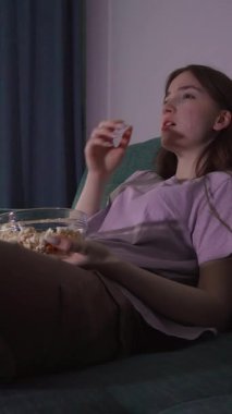 Genç kız kanepeye oturup film izleyerek loş ışıklı oda dikey videosunda cam kaseden patlamış mısır yemekten zevk alıyor.