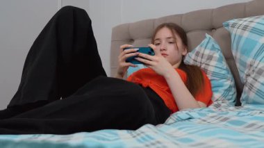 Genç kız gündelik giysiler içinde yatakta dinleniyor, neşeli bir ifadeyle akıllı telefonlara bakıyor evde sakin bir şekilde eğleniyor ve akıllı telefondan oyun oynuyor..