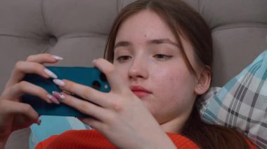 Genç kız gündelik giysiler içinde yatakta dinleniyor, neşeli bir ifadeyle akıllı telefonlara bakıyor evde sakin bir şekilde eğleniyor ve akıllı telefondan oyun oynuyor..