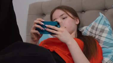 Genç kız gündelik giysiler içinde yatakta dinleniyor, neşeli bir ifadeyle akıllı telefonlara bakıyor evde sakin bir şekilde eğleniyor ve akıllı telefondan oyun oynuyor..
