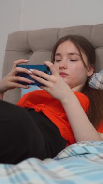 Genç kız gündelik giysiler içinde yatakta dinleniyor, neşeli bir ifadeyle akıllı telefonuna göz atıyor evde boş vakitlerin tadını çıkarıyor ve akıllı telefondan oyun oynuyor. dikey video