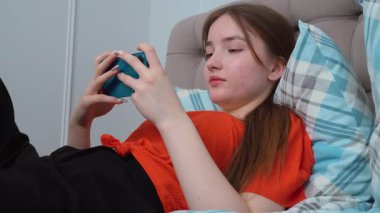 Rahat genç kız akıllı telefonuyla oyun oynuyor rahat giysiler içinde yatakta uzanıyor ve evde kişisel rahatlama anlarının tadını çıkarıyor.
