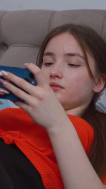 Genç kız gündelik giysiler içinde yatakta dinleniyor, neşeli bir ifadeyle akıllı telefonuna göz atıyor evde boş vakitlerin tadını çıkarıyor ve akıllı telefondan oyun oynuyor. dikey video