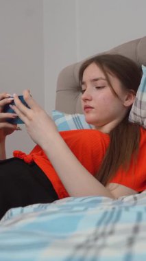Rahat genç kız akıllı telefonuyla oyun oynuyor rahat giysiler içinde yatağında uzanıyor ve evde dikey video kasetlerde dinleniyor.