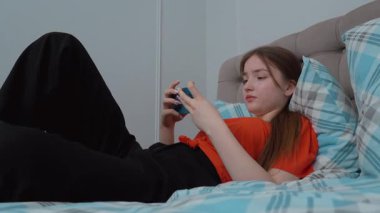 Rahat genç kız akıllı telefonuyla oyun oynuyor rahat giysiler içinde yatakta uzanıyor ve evde kişisel rahatlama anlarının tadını çıkarıyor.