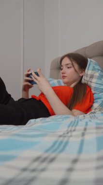 Rahat genç kız akıllı telefonuyla oyun oynuyor rahat giysiler içinde yatağında uzanıyor ve evde dikey video kasetlerde dinleniyor.