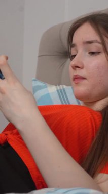 Genç kız rahat bir şekilde yatakta uzanıyor, günlük turuncu gömlekler giyiyor ve akıllı telefonuyla sosyal medyada geziniyor ve sakin bir yatak odasında dikey video ortamında rahatlarken aksiyon oyunları oynuyor.