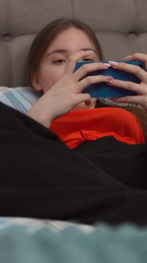 Genç kız rahat bir şekilde yatakta uzanıyor, günlük turuncu gömlekler giyiyor ve akıllı telefonuyla sosyal medyada geziniyor ve sakin bir yatak odasında dikey video ortamında rahatlarken aksiyon oyunları oynuyor.