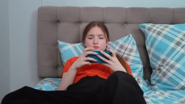 Genç kız rahat bir şekilde yatakta uzanıyor, günlük turuncu gömlekler giyip sosyal medyada akıllı telefonuyla geziniyor ve sakin bir yatak odasında dinlenirken aksiyon oyunları oynuyor.