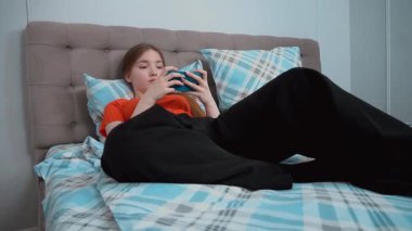 Rahatlamış genç kız yatakta rahat bir şekilde uzanıyor, akıllı telefon ekranlarına bakıyor, oyun oynuyor, sosyal medyadan ve hareketli oyunlardan keyif alıyor.