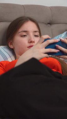 Rahat genç kız siyah battaniyenin altında uzanmış mavi akıllı telefon kullanıyor, mobil oyunlar oynuyor ya da sosyal medyada geziniyor, ekran dikey video izliyor.