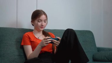 Rahat oturma odasındaki yeşil kanepede dinlenirken gezici oyunların keyfini çıkaran günlük kıyafetli genç kız akıllı telefon aracılığıyla dijital eğlencenin tadını çıkarıyor.