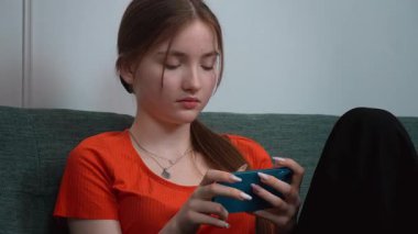 Rahat oturma odasındaki yeşil kanepede dinlenirken gezici oyunların keyfini çıkaran günlük kıyafetli genç kız akıllı telefon aracılığıyla dijital eğlencenin tadını çıkarıyor.