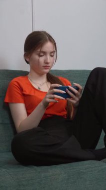 Rahat oturma odasındaki yeşil kanepede dinlenirken hareketli oyunlardan hoşlanan genç kız akıllı telefonlu dikey video ile dijital eğlencenin tadını çıkarıyor.