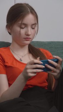 Rahat oturma odasındaki yeşil kanepede dinlenirken hareketli oyunlardan hoşlanan genç kız akıllı telefonlu dikey video ile dijital eğlencenin tadını çıkarıyor.