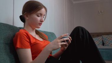 Rahat oturma odasındaki yeşil kanepede dinlenirken gezici oyunların keyfini çıkaran günlük kıyafetli genç kız akıllı telefon aracılığıyla dijital eğlencenin tadını çıkarıyor.