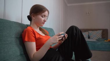 Rahat oturma odasındaki yeşil kanepede dinlenirken gezici oyunların keyfini çıkaran günlük kıyafetli genç kız akıllı telefon aracılığıyla dijital eğlencenin tadını çıkarıyor.