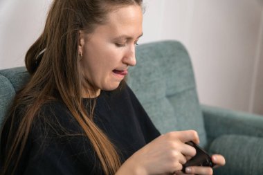 Uzun kahverengi saçlı, siyah tişörtlü, rahat bir koltukta oturan, hareketli bir oyun oynarken dudağını ısıran genç bir kadın.