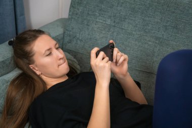 Uzun kahverengi saçlı, siyah tişörtlü genç bir kadın rahat koltukta uzanıyor, akıllı telefon kullanıyor, iki eliyle yatay olarak tutuyor ve odaklanmış bir ifadeyle ekrana bakıyor.