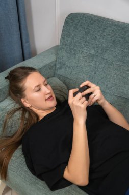 Genç kadın rahat bir kanepede uzanıyor ve akıllı telefon kullanıyor, mobil oyunlar oynuyor, internette geziniyor, haberleri okuyor, sosyal ağlarda sohbet ediyor veya çevrimiçi alışveriş yapıyor. Dikey fotoğraf