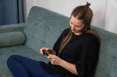 Genç bir kadın oturma odasındaki rahat bir koltukta dinlenirken akıllı telefon kullanıyor, mobil teknolojinin ve günlük ev ortamında internet iletişiminin tadını çıkarıyor.