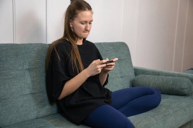 Genç bir kadın modern oturma odasındaki bir kanepede rahatça oturuyor, evinde boş zamanlarında cep telefonuyla gezintiye çıkıyor.