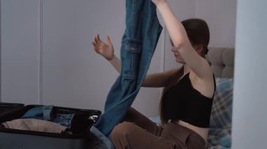 Genç bir kadın, giysilerini düzenli olarak katlıyor ve kişisel giysilerini seyahat çantasına yerleştiriyor. Yatakta oturuyor ve yaklaşan yolculuğa hazırlanıyor.