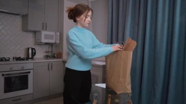 Genç bir kız, şık ve modern bir mutfakta, kese kağıdından taze ürünler ve diğer yiyecek eşyaları çıkarıyor, sağlıklı yaşam tarzını ve ev hayatını ön plana çıkarıyor.