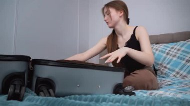 Genç bir kadın, giysilerini düzenli olarak katlıyor ve kişisel giysilerini seyahat çantasına yerleştiriyor. Yatakta oturuyor ve yaklaşan yolculuğa hazırlanıyor.